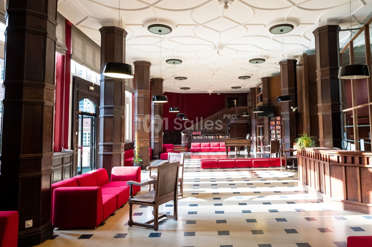 Salle lumineuse avec colonnes en bois, canapés rouges, sol en damier et escalier menant à une mezzanine.