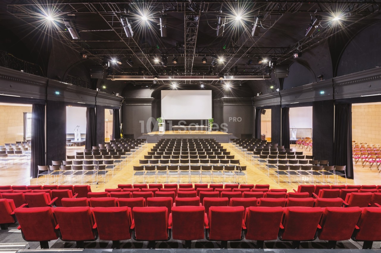 Salle de conférence avec des rangées de sièges rouges et noirs, une scène équipée d'un écran blanc et un éclairage au…