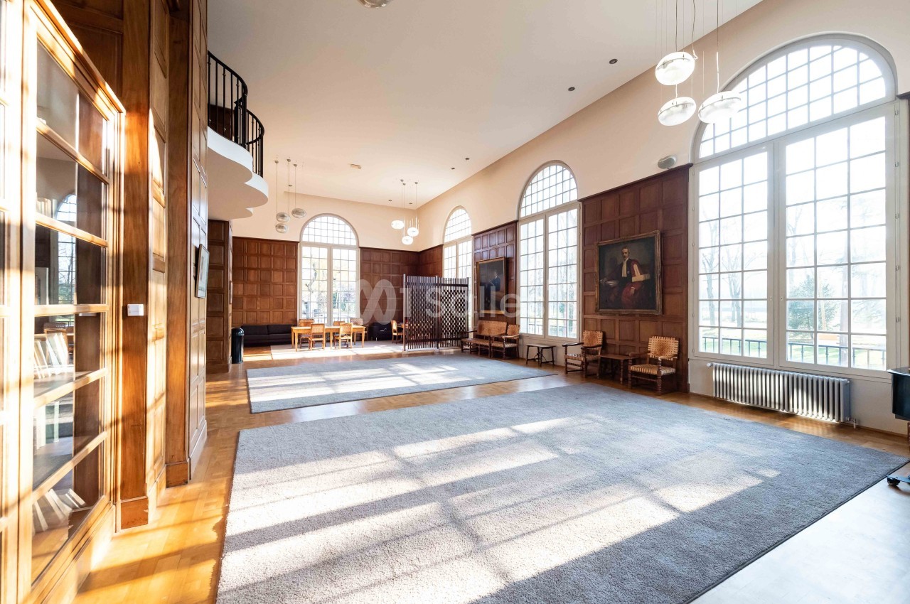 Salle lumineuse avec de grandes fenêtres cintrées, parquet en bois, tapis gris et mobilier classique en bois.