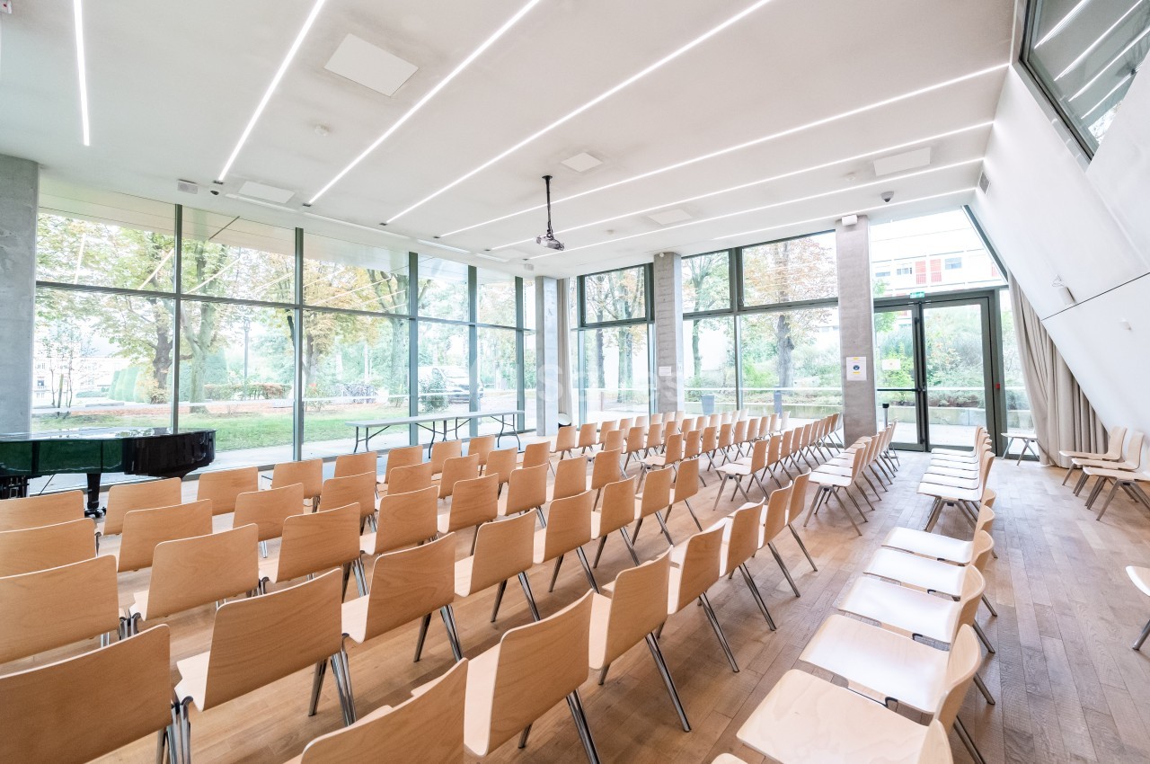Salle lumineuse avec des rangées de chaises en bois, grandes baies vitrées donnant sur un espace arboré.