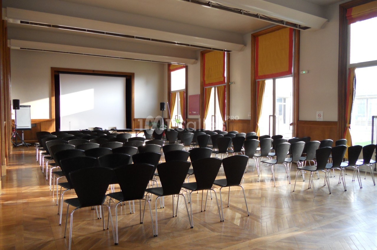Salle de conférence lumineuse avec des rangées de chaises noires face à un écran de projection.