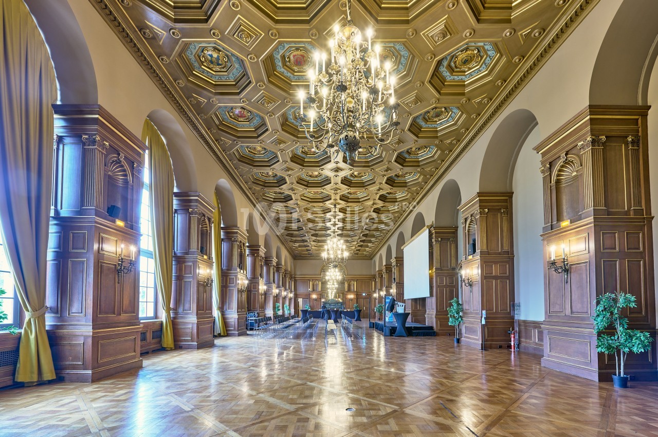 Grande salle avec parquet, hauts plafonds ornés, lustres imposants, colonnes en bois et rideaux encadrant des fenêtres.