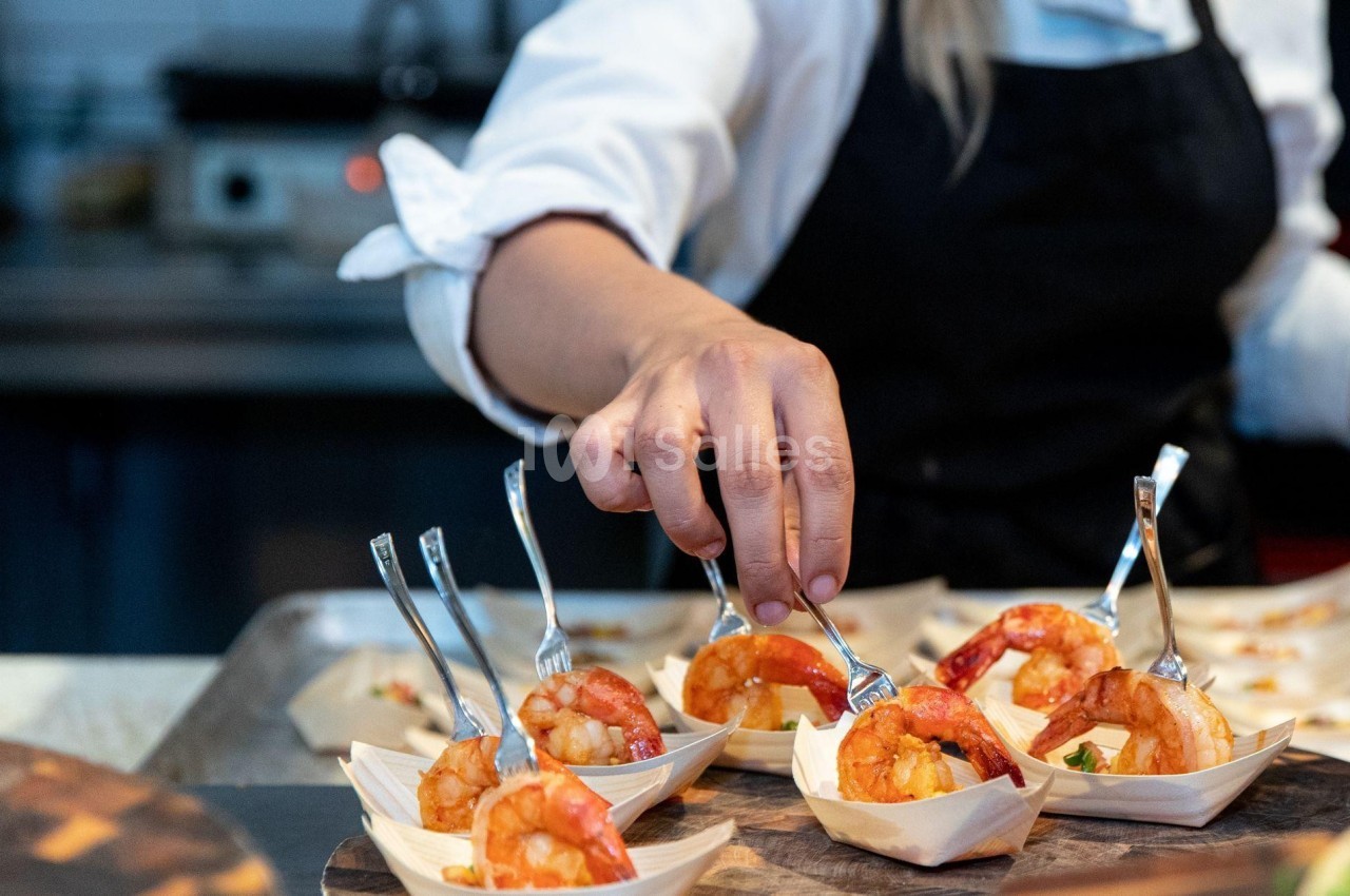 Une main dispose des crevettes dans des coupelles en bois avec des fourchettes, dans une cuisine professionnelle.