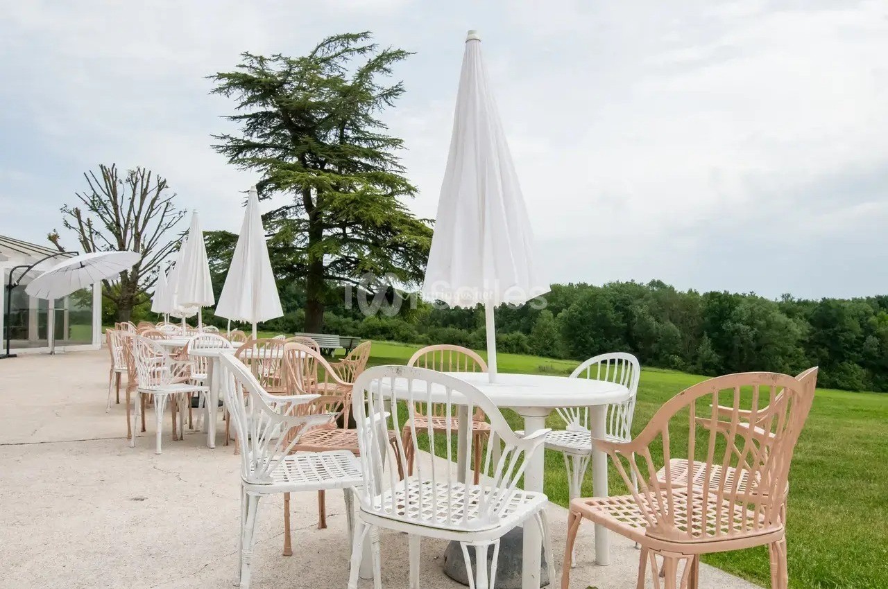 Terrasse avec tables et chaises blanches et roses, parasols ouverts, donnant sur un espace vert arboré.