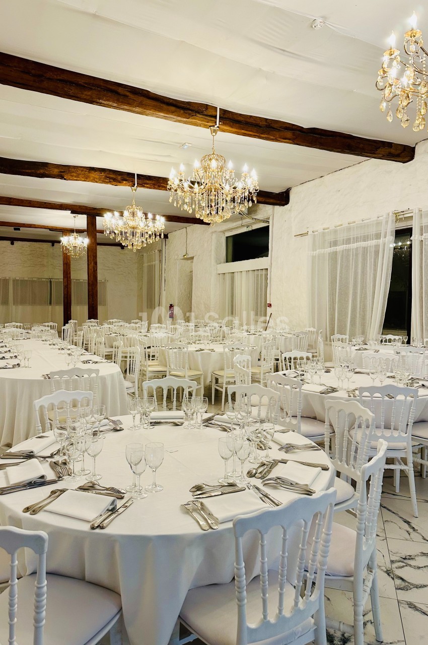 Salle de réception élégante avec tables rondes dressées, chaises blanches et lustres en cristal suspendus.