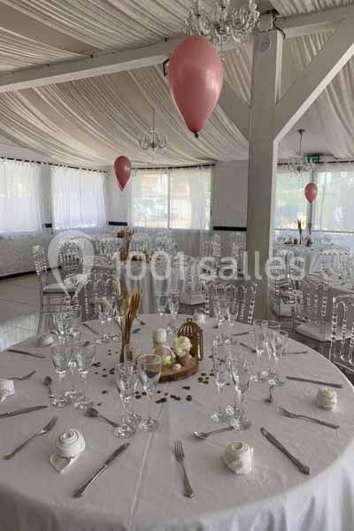 Salle de réception décorée avec des tables rondes dressées, nappes blanches, ballons roses et lumière naturelle.