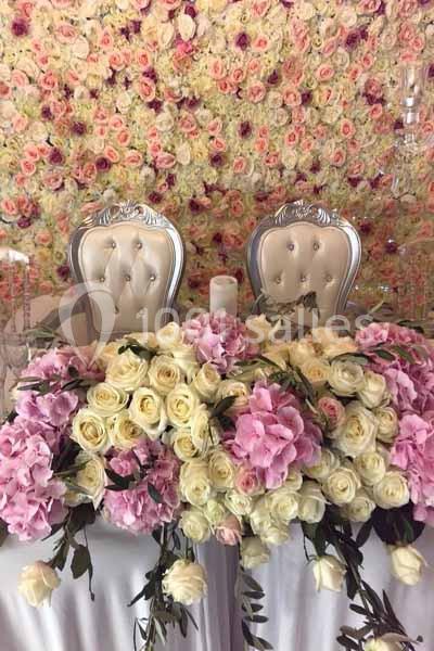 Deux chaises argentées devant une table ornée de roses blanches et roses, avec un mur fleuri en arrière-plan.