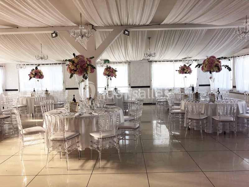 Salle de réception élégante avec tables rondes dressées, chaises transparentes et décorations florales.