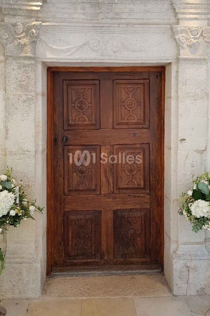 Porte en bois sculptée encadrée de pierre, décorée de deux arrangements floraux sur des supports métalliques.