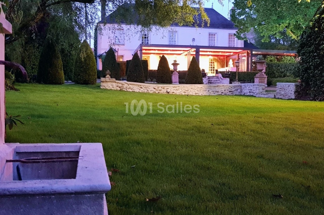 Jardin verdoyant au crépuscule avec une maison éclairée en arrière-plan et un bassin en pierre au premier plan.
