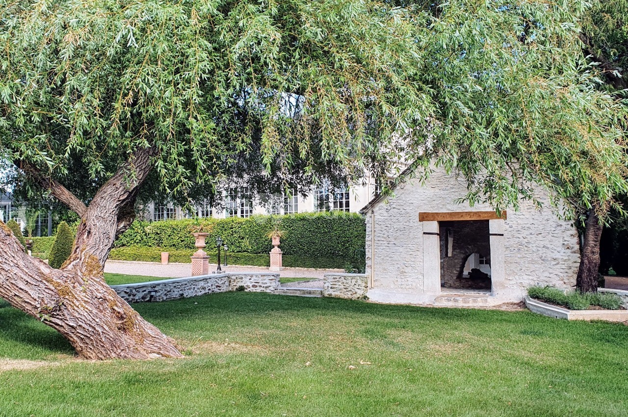 Petit bâtiment en pierre entouré d'herbe, d'arbres et de haies dans un jardin bien entretenu.