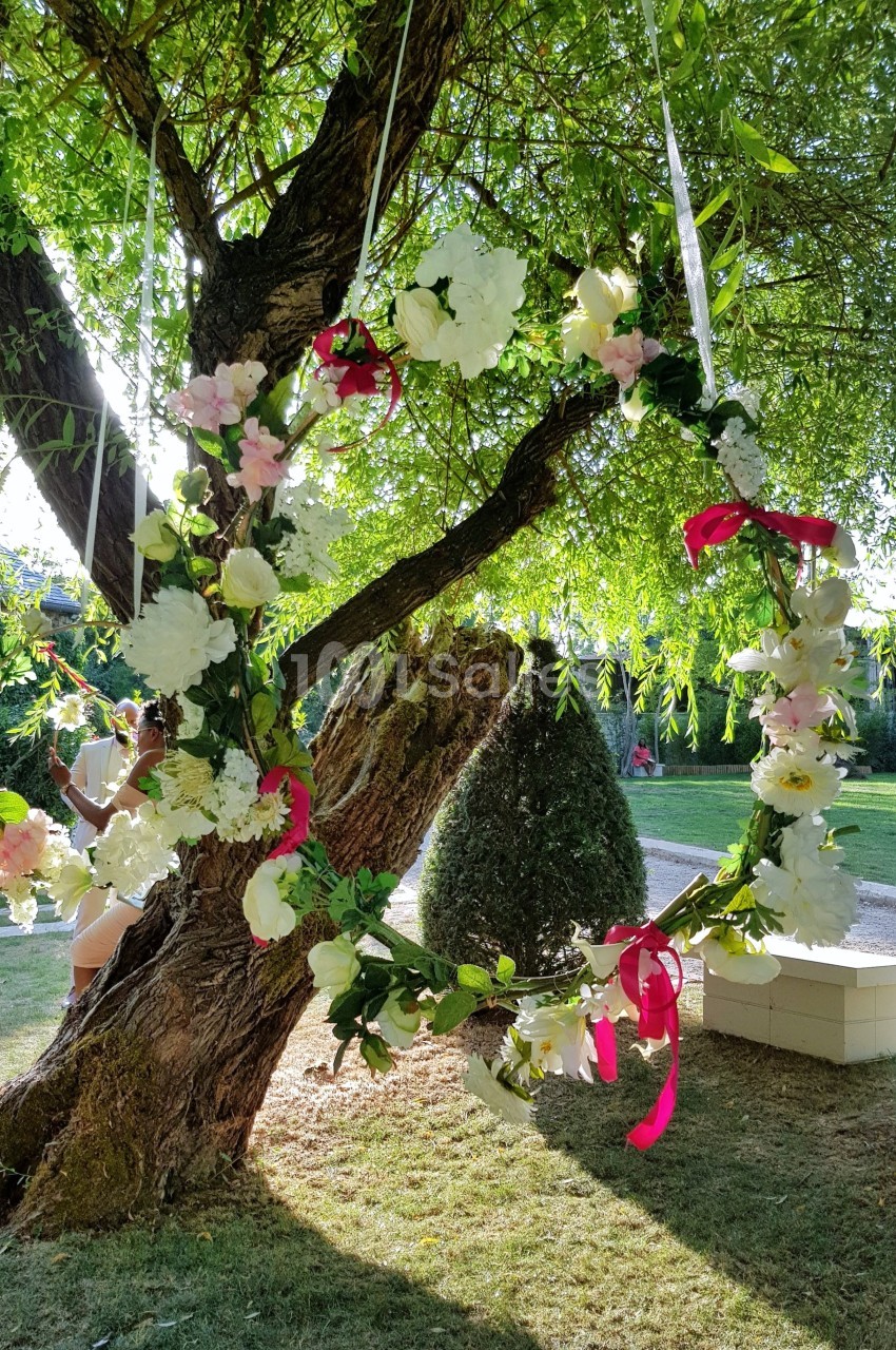 Couronne de fleurs blanches et roses suspendue à un arbre sous la lumière du soleil dans un jardin verdoyant.