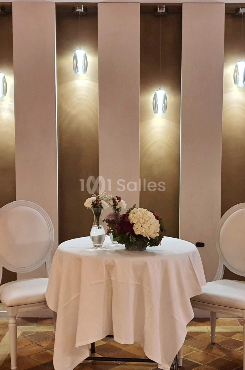 Table ronde avec nappe blanche, deux chaises blanches et un bouquet de fleurs, éclairée par des appliques murales.