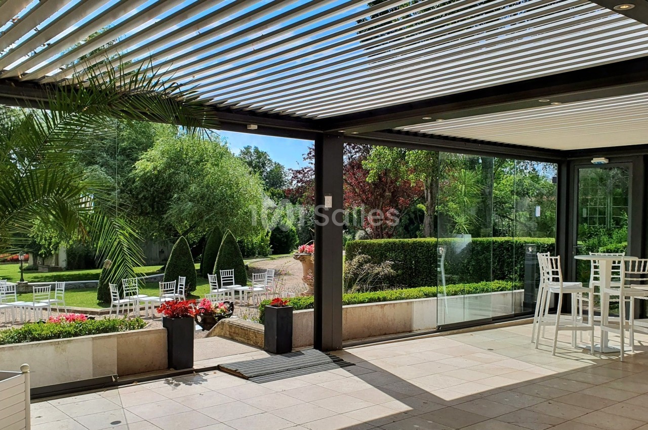 Terrasse couverte avec pergola à lames orientables, vue sur un jardin arboré et mobilier de jardin blanc.