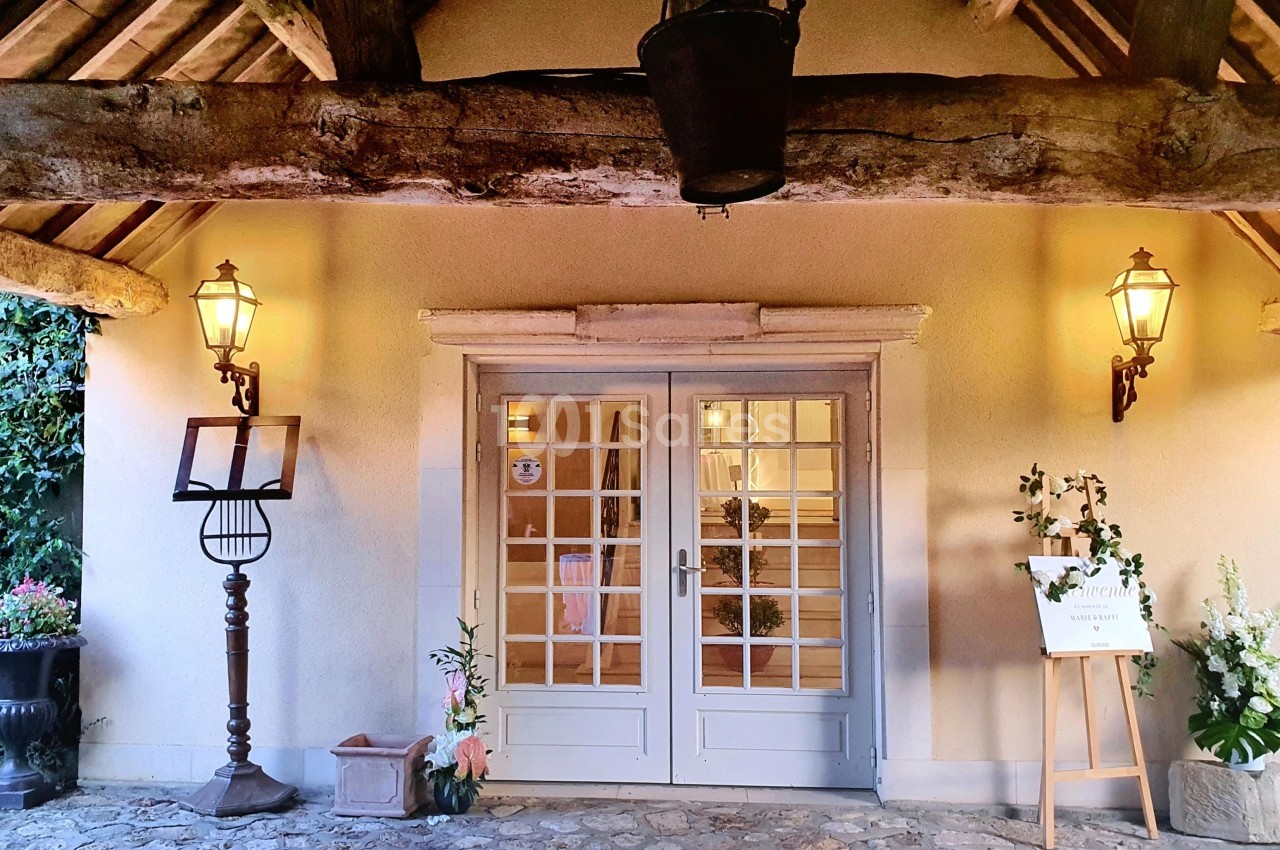 Entrée d'un bâtiment avec porte vitrée, encadrée de deux lanternes murales et décorée de plantes et d'un chevalet.