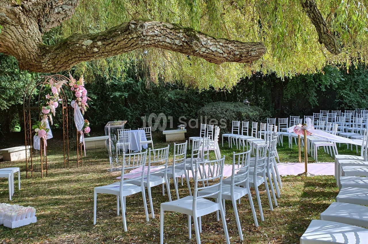 Chaises blanches disposées en extérieur sous un arbre, avec une arche fleurie et une allée décorée pour une cérémonie.