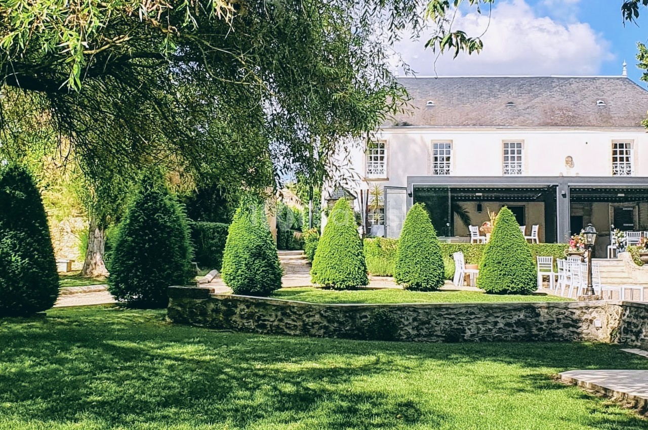 Jardin verdoyant avec pelouse, arbres et arbustes taillés, devant une maison blanche à toit d'ardoise.