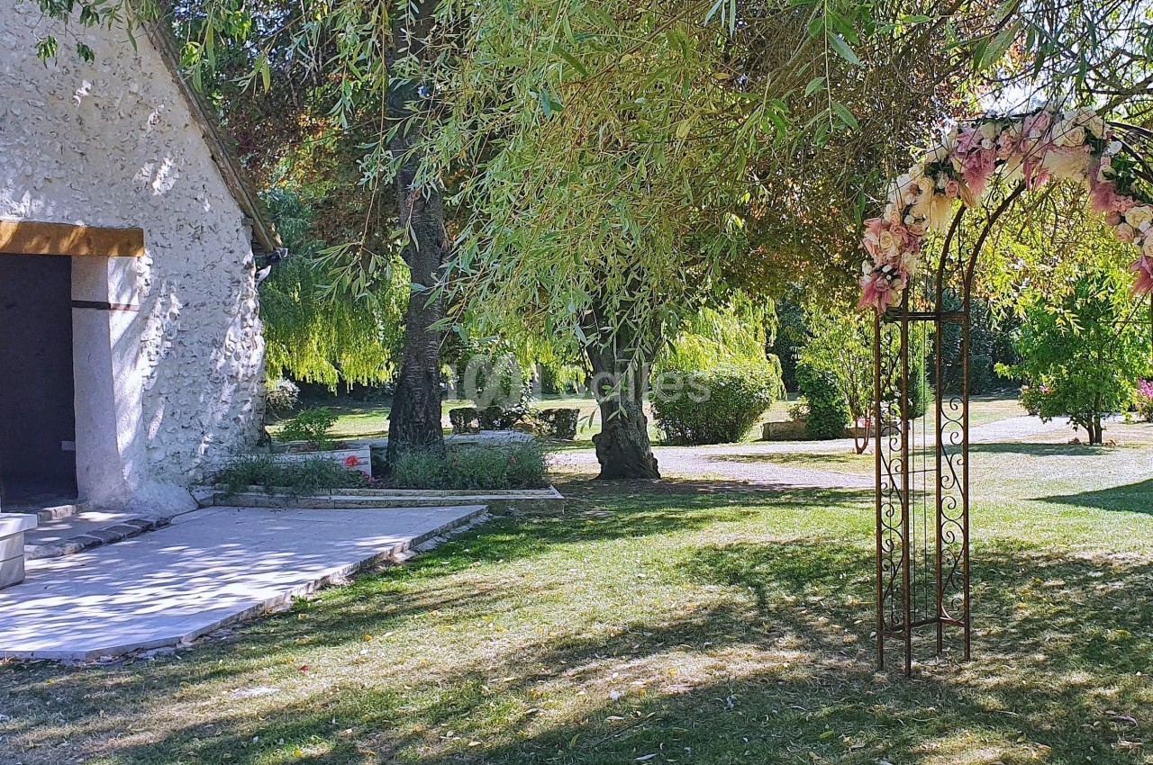 Arche fleurie en métal dans un jardin verdoyant, près d'un bâtiment en pierre avec une porte ouverte.