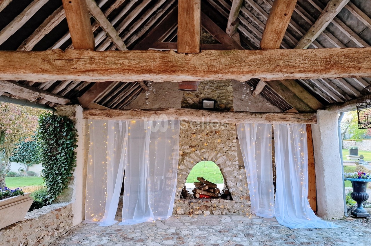 Structure en pierre avec toit en bois, rideaux blancs lumineux et ouverture centrale décorée de bûches.