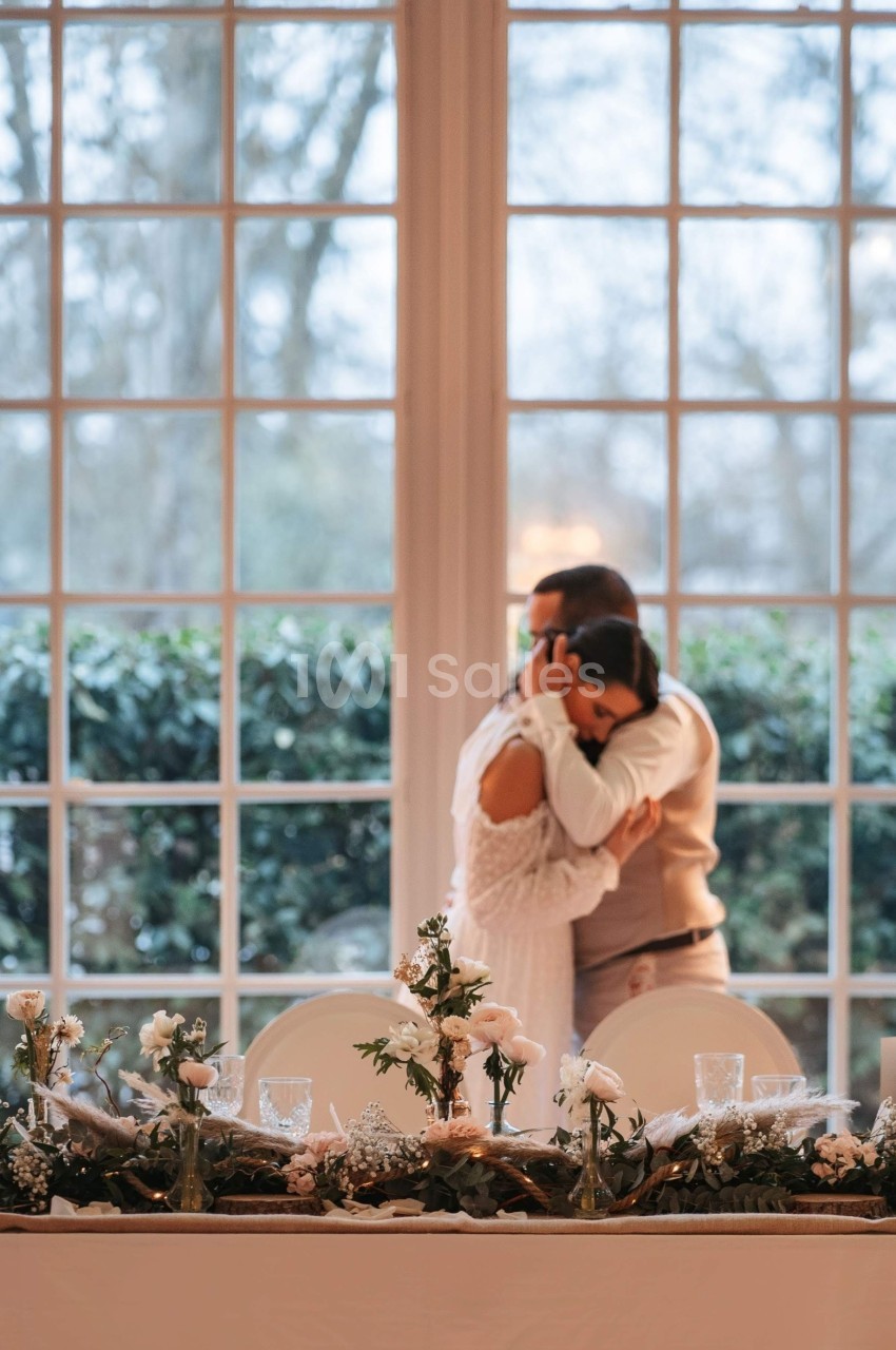 Un couple s'enlace devant une grande baie vitrée, avec une table décorée de fleurs et de feuillages au premier plan.