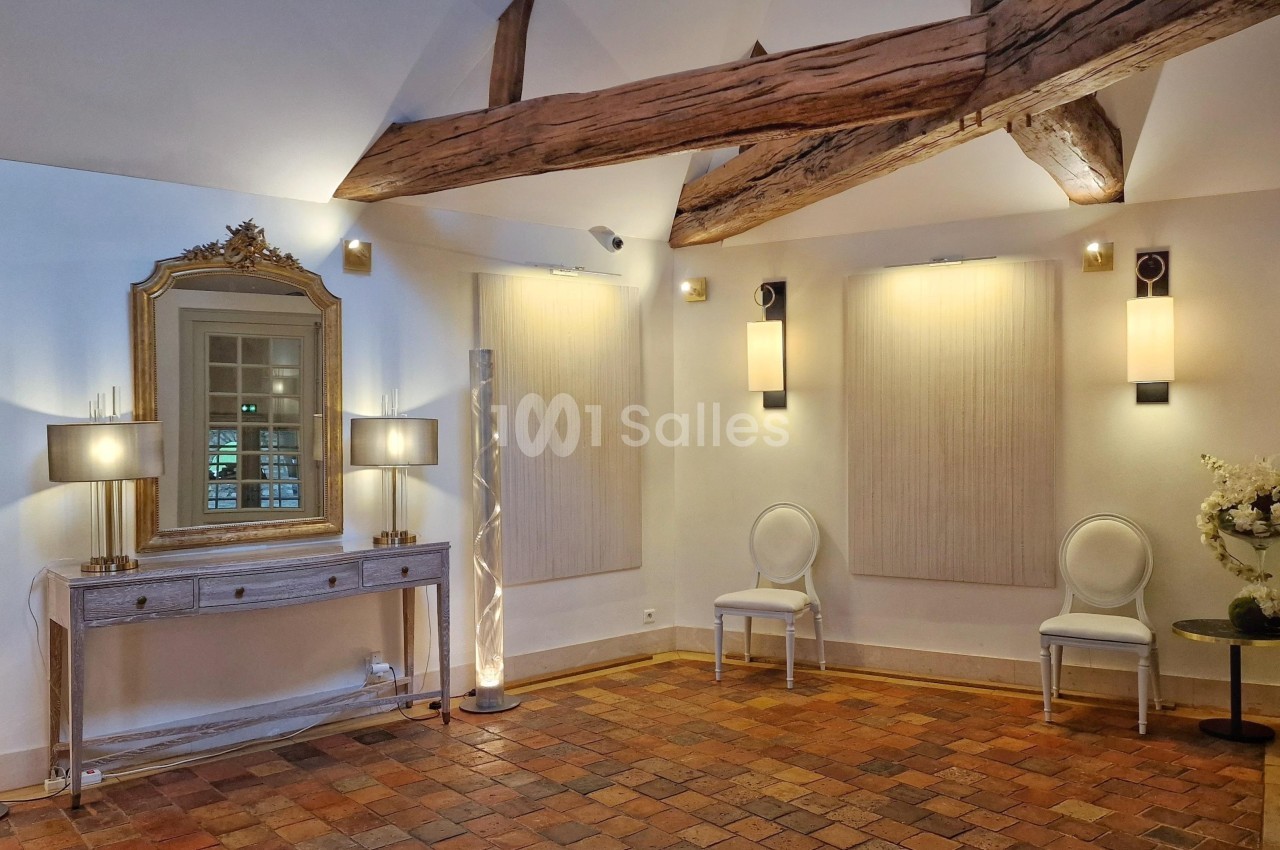 Salle lumineuse avec poutres apparentes, sol en terre cuite, console avec miroir doré et chaises blanches.