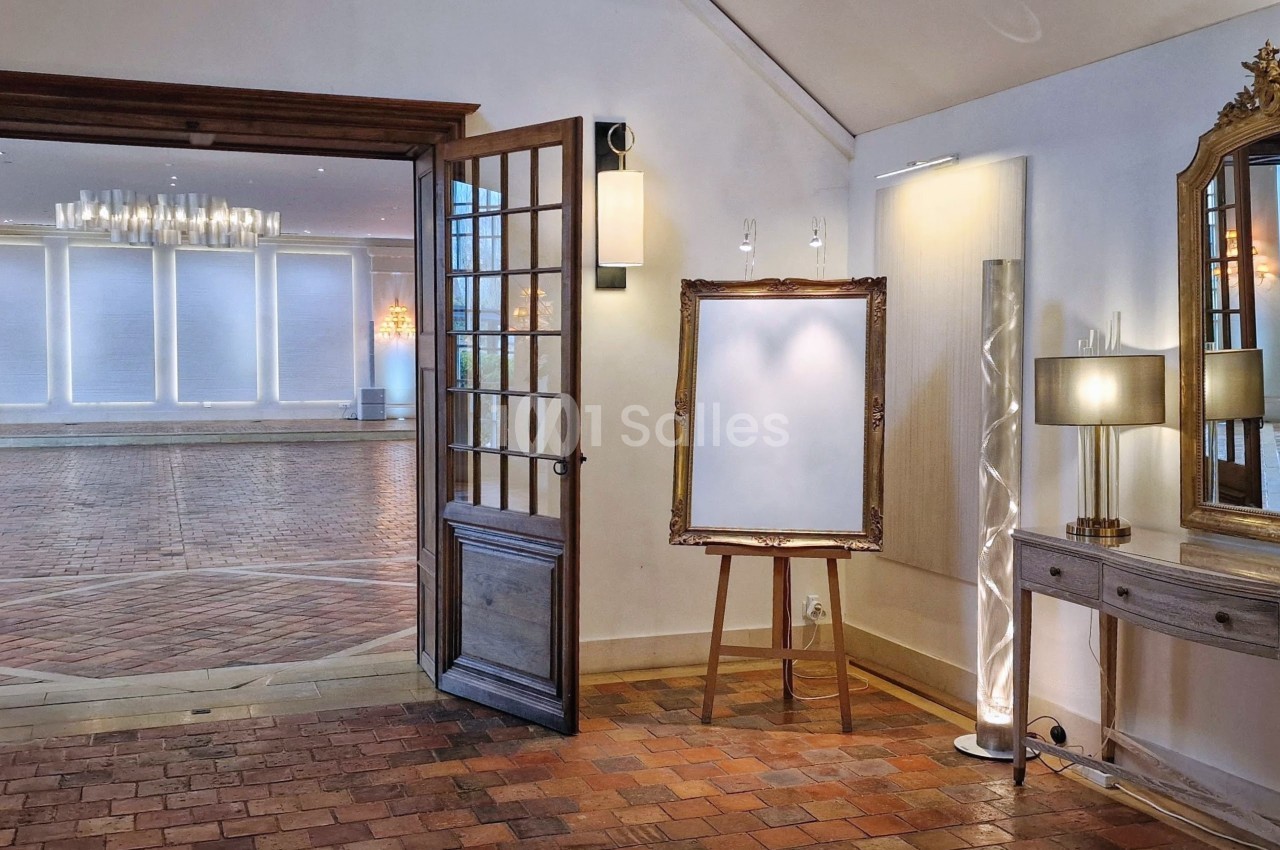 Salle lumineuse avec portes vitrées ouvertes, chevalet vide, console avec miroir doré et lampes, sol en briques.