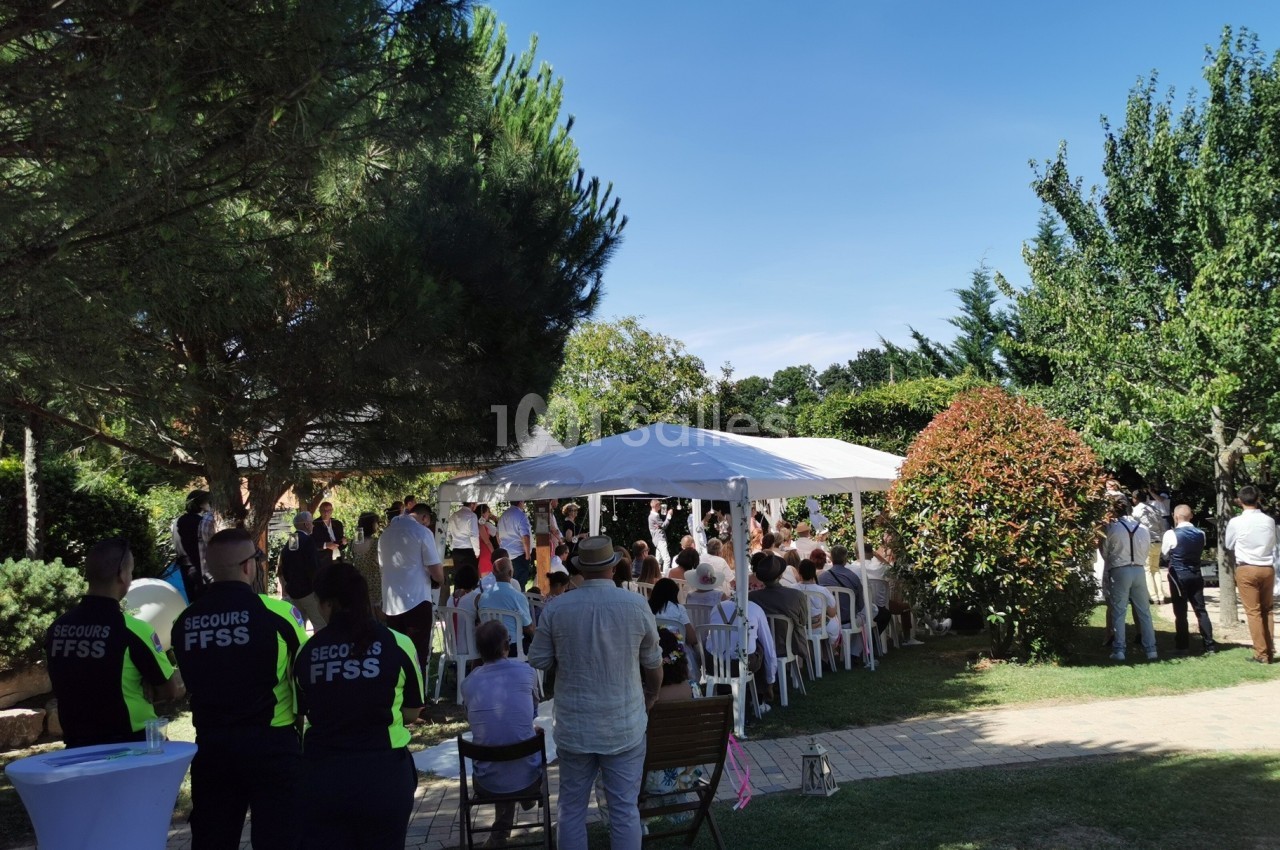 Groupe de personnes assises sous un chapiteau blanc lors d'un événement en extérieur, entouré d'arbres et de verdure.