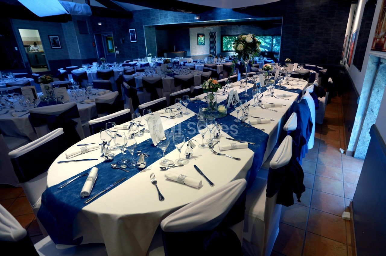 Salle de réception décorée avec des tables dressées, nappes blanches et chemins de table bleus, prête pour un événement.
