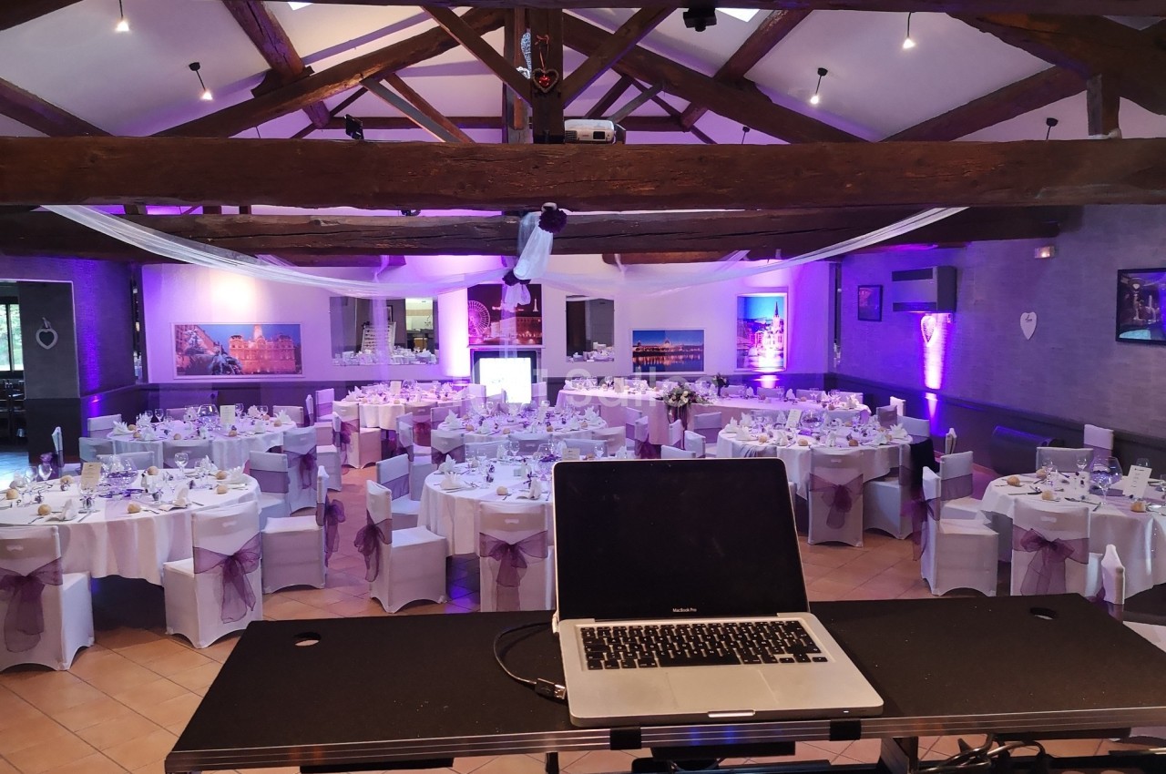 Salle de réception décorée pour un événement, avec tables dressées et éclairages violets, vue depuis une table de DJ.
