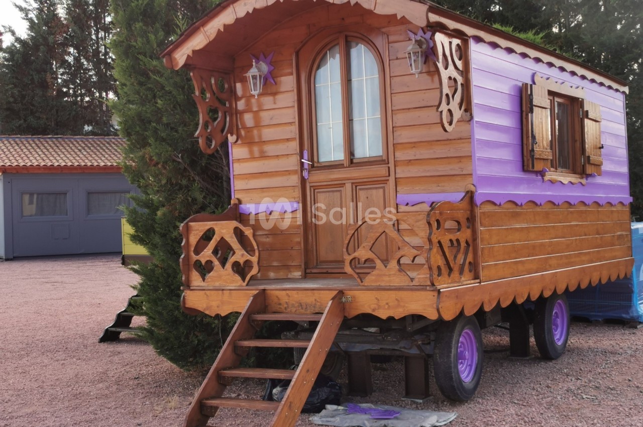 Roulotte en bois et violet avec des décorations sculptées, posée sur roues, entourée d'arbres et de bâtiments.