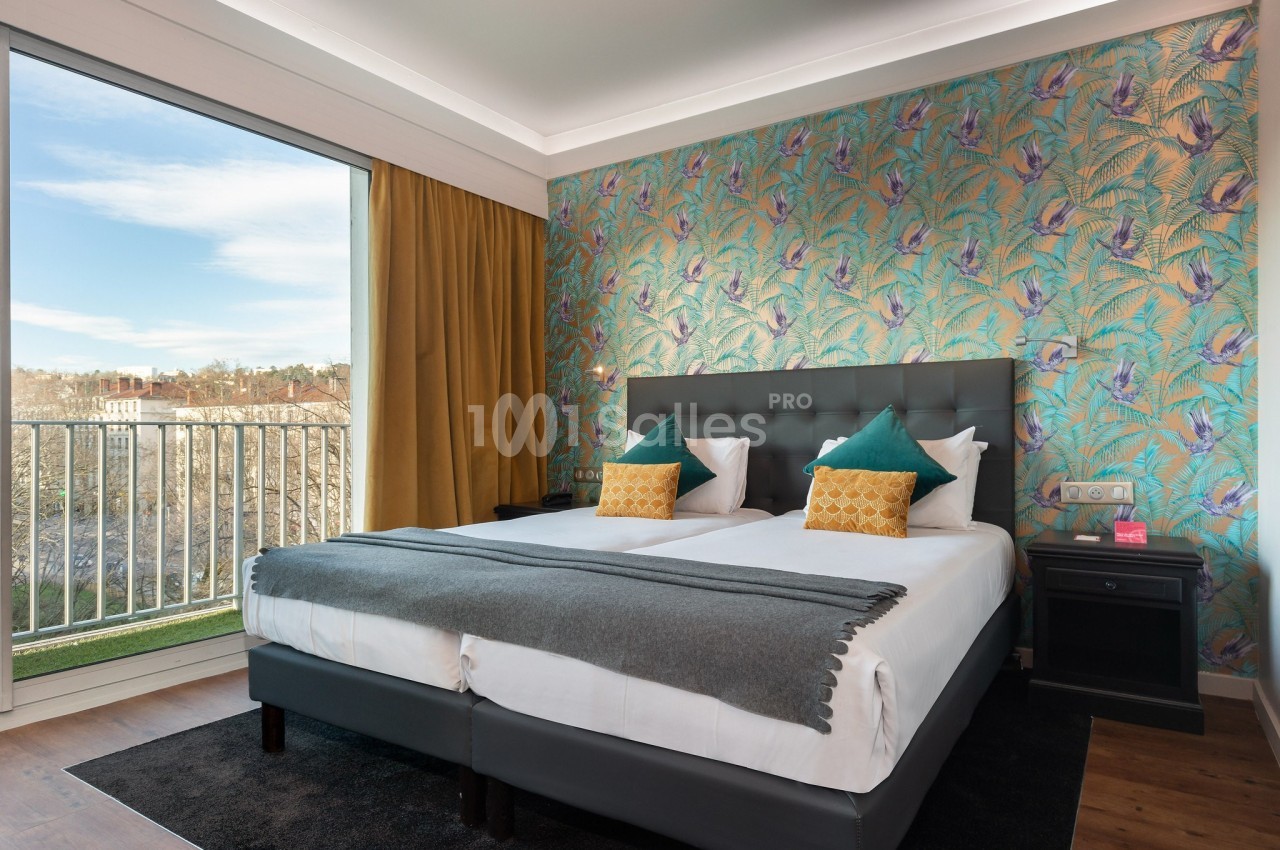 Chambre lumineuse avec lit double, coussins colorés, mur à motifs tropicaux et grande fenêtre donnant sur l'extérieur.