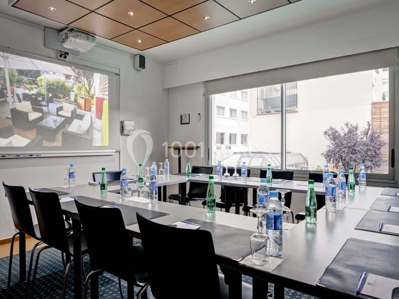 Salle de réunion lumineuse avec table rectangulaire, chaises, bouteilles d'eau, écran de projection et vue sur l'extérieur.