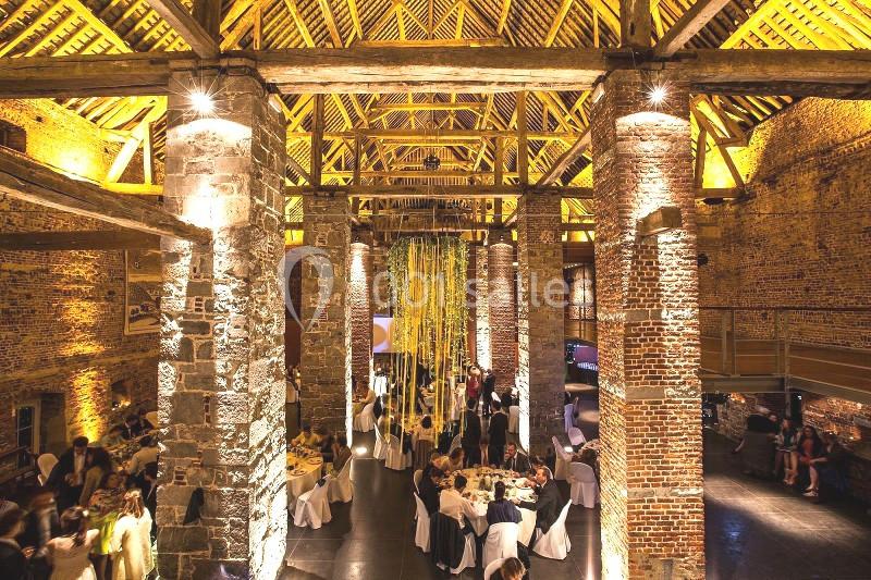 Salle en pierre et bois avec plafond voûté, éclairage chaleureux, tables dressées et invités lors d'un événement.