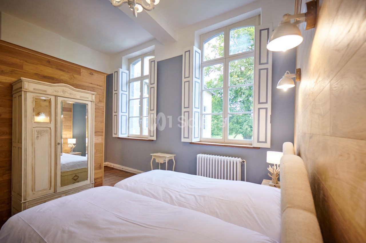 Chambre lumineuse avec deux lits simples, armoire ancienne, murs en bois et fenêtres donnant sur un jardin.