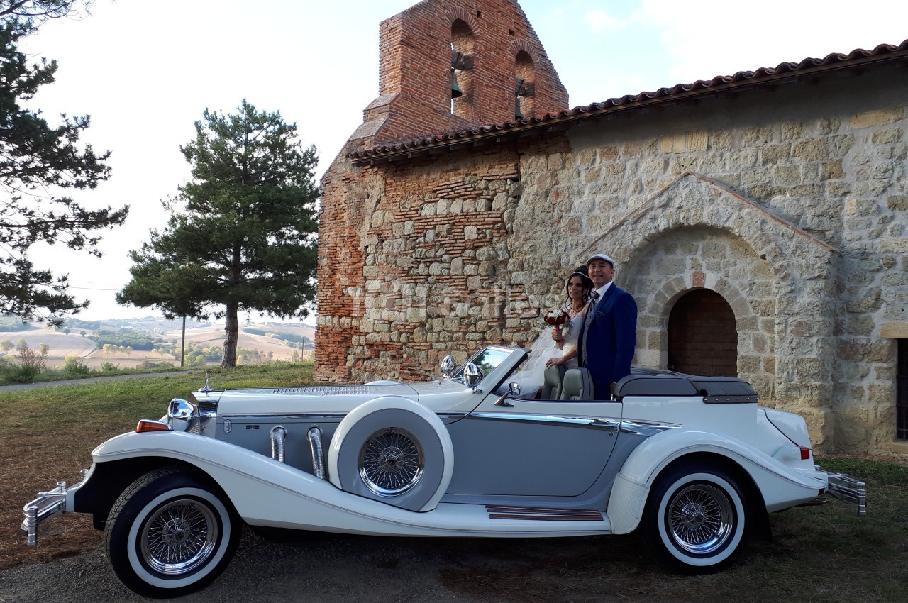 Un couple en tenue de mariage pose près d'une voiture ancienne blanche et grise devant une église en pierre.