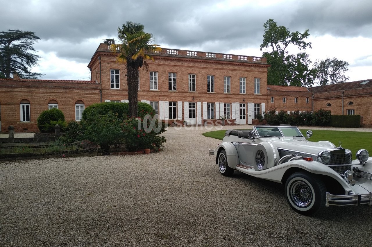 Voiture ancienne blanche garée devant un grand bâtiment en briques rouges entouré de végétation.