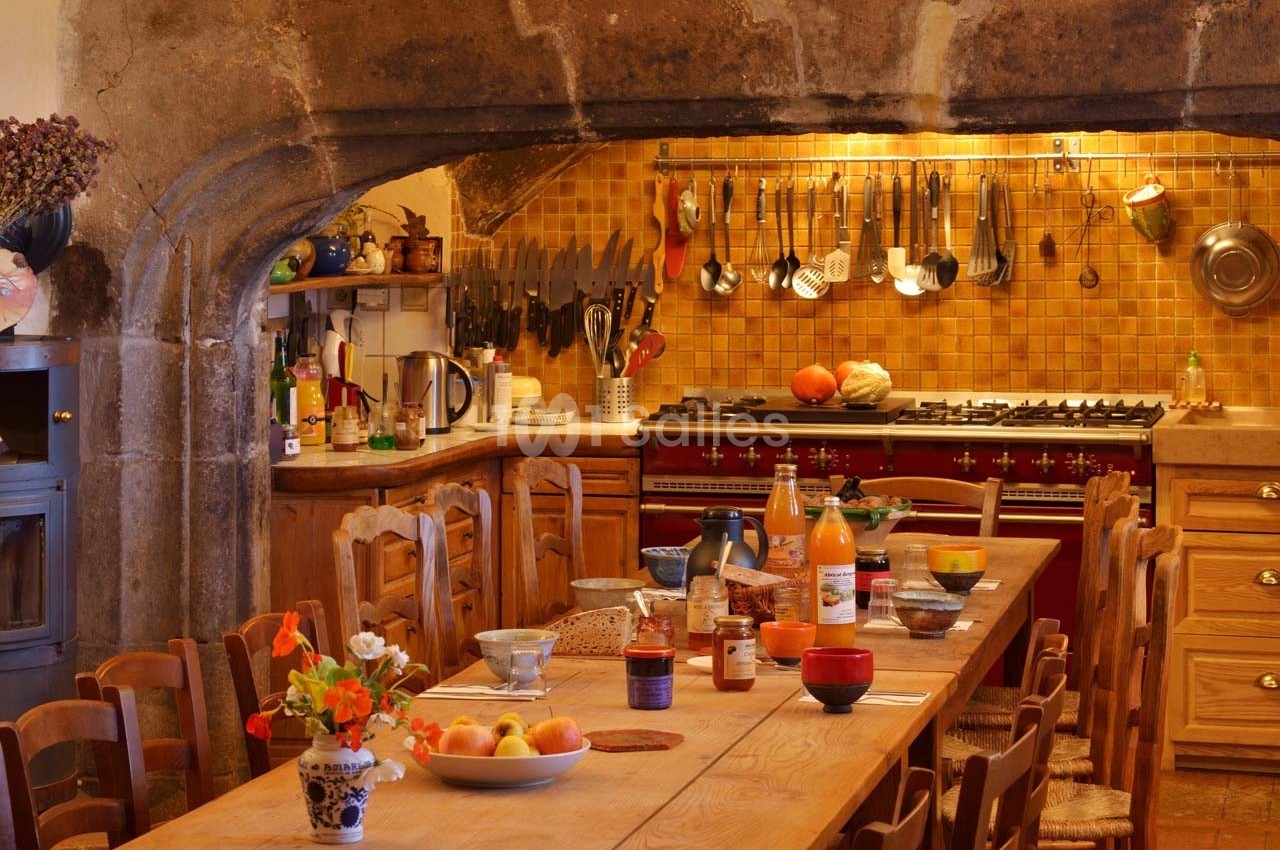 Cuisine rustique avec une grande table en bois dressée, des ustensiles accrochés et un fourneau sous une hotte en pierre.