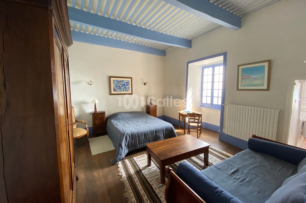 Chambre lumineuse avec lit double, canapé, meubles en bois, plafond avec poutres bleues et fenêtre donnant sur l'extérieur.