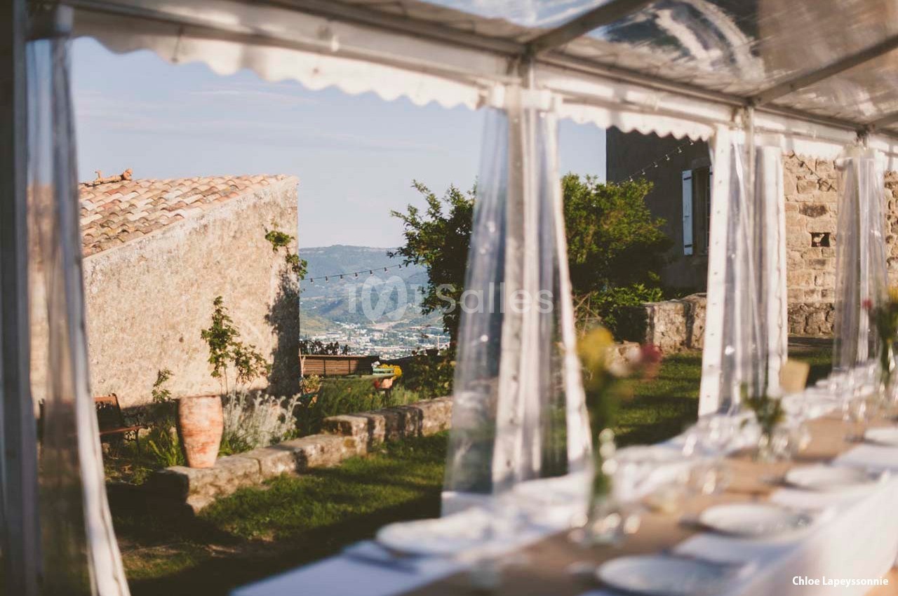 Table dressée sous une tente transparente avec vue sur un jardin, des collines et des bâtiments en pierre.
