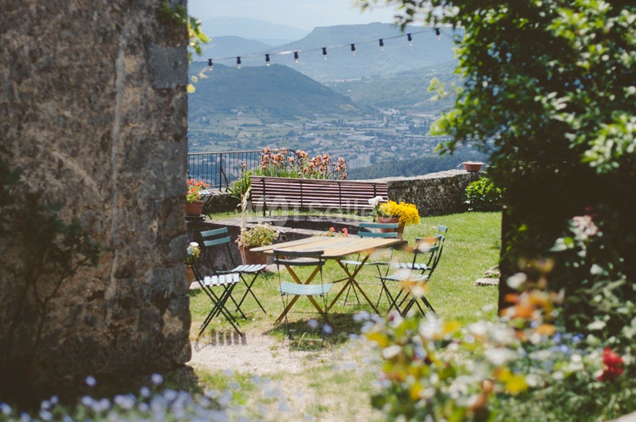 Table et chaises colorées dans un jardin fleuri avec vue sur des collines et guirlande lumineuse suspendue.