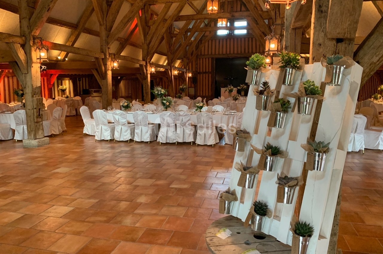 Salle de réception avec poutres apparentes, tables rondes dressées et un présentoir de plantes en pots au premier plan.