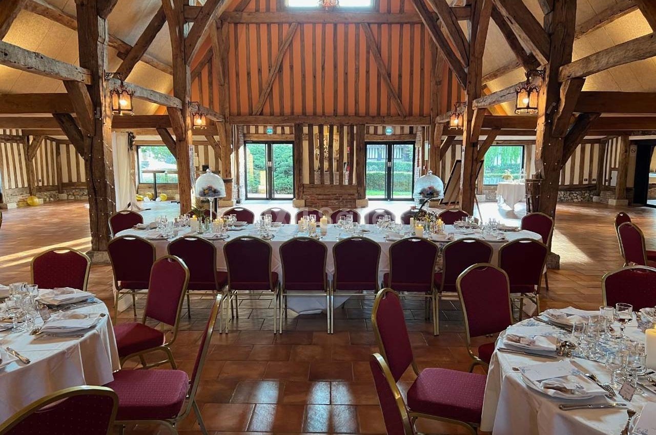 Salle de réception avec charpente en bois, tables rondes dressées avec nappes blanches et chaises rouges.