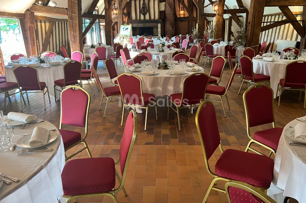 Salle de réception avec tables rondes dressées, nappes blanches et chaises rouges, dans un cadre rustique avec poutres…