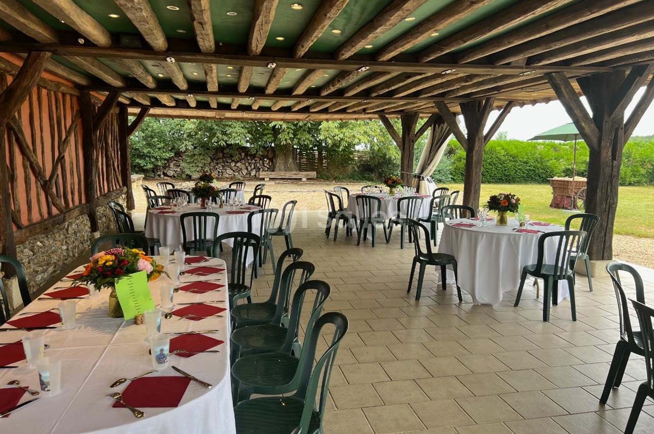 Tables rondes dressées avec nappes blanches et chaises vertes sous une pergola en bois, donnant sur un jardin.
