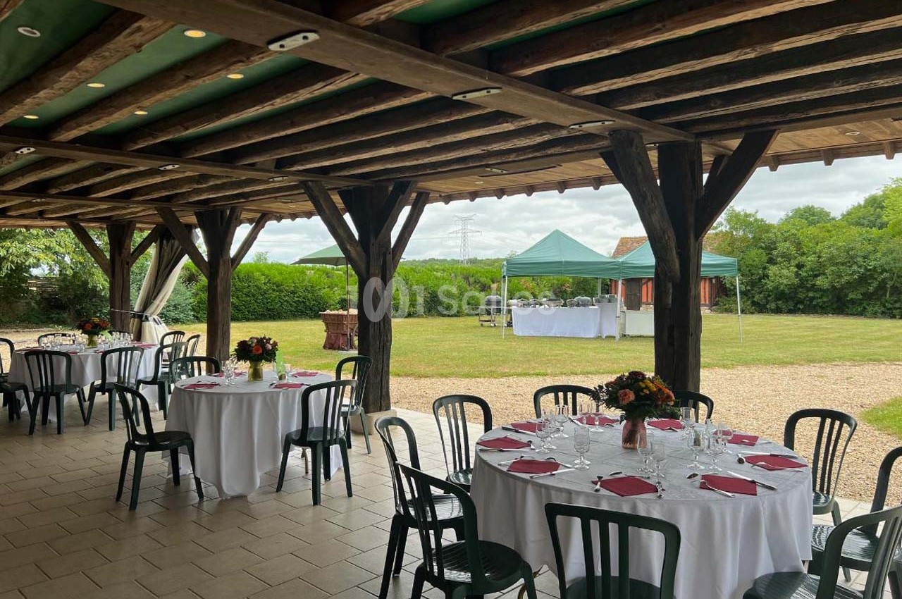 Salle couverte avec tables dressées, vue sur un jardin et une tente extérieure pour un événement en plein air.