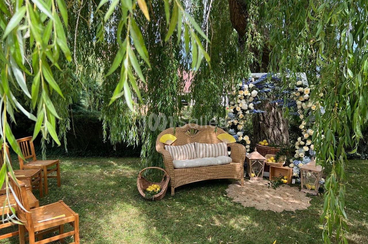 Espace extérieur décoré avec un canapé en osier, des fleurs, des chaises en bois et des branches de saule tombantes.