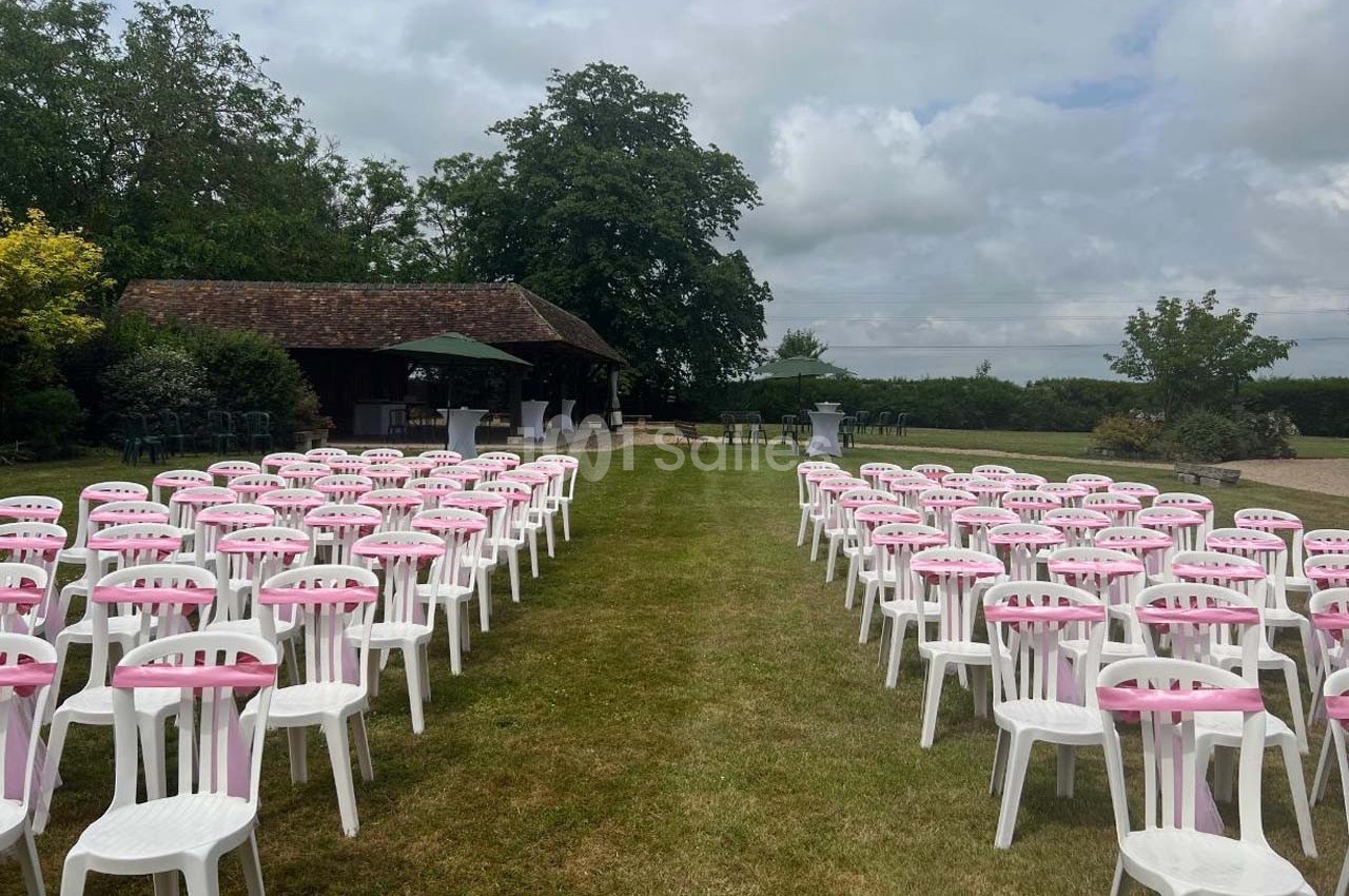 Chaises blanches avec housses roses disposées en rangées sur une pelouse, face à une petite structure en bois.