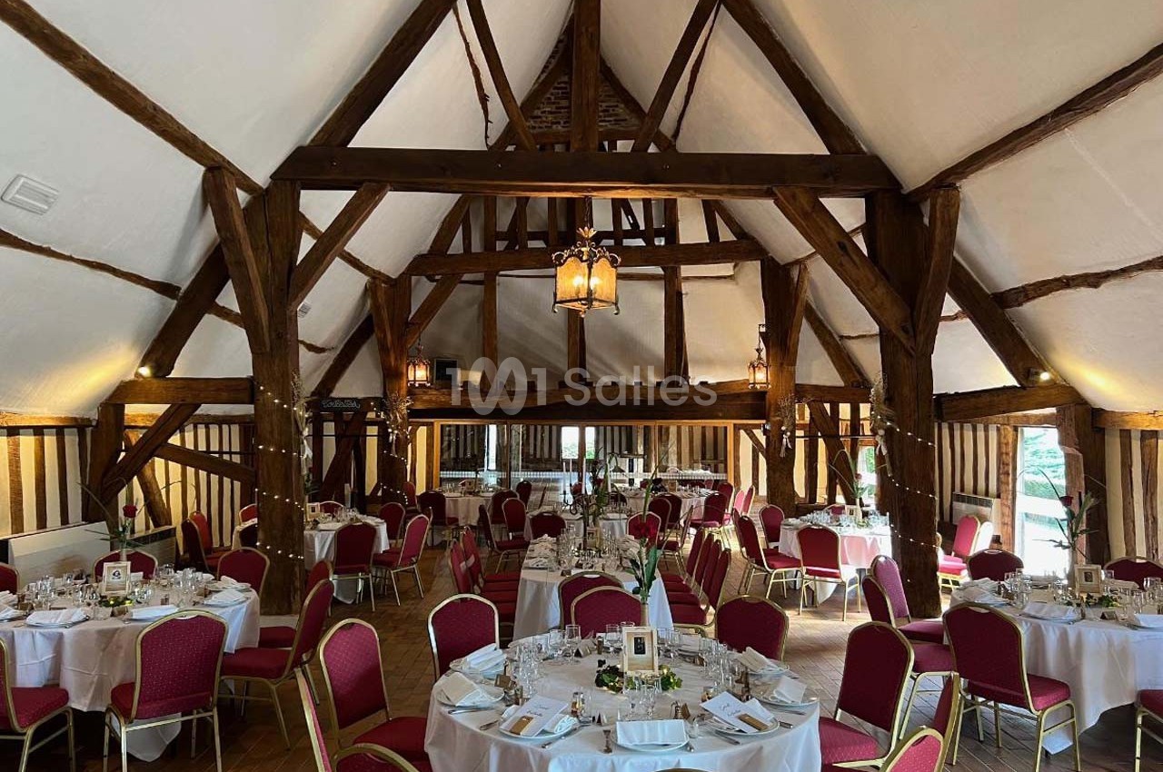 Salle de réception rustique avec poutres en bois apparentes, tables rondes dressées et chaises rouges.