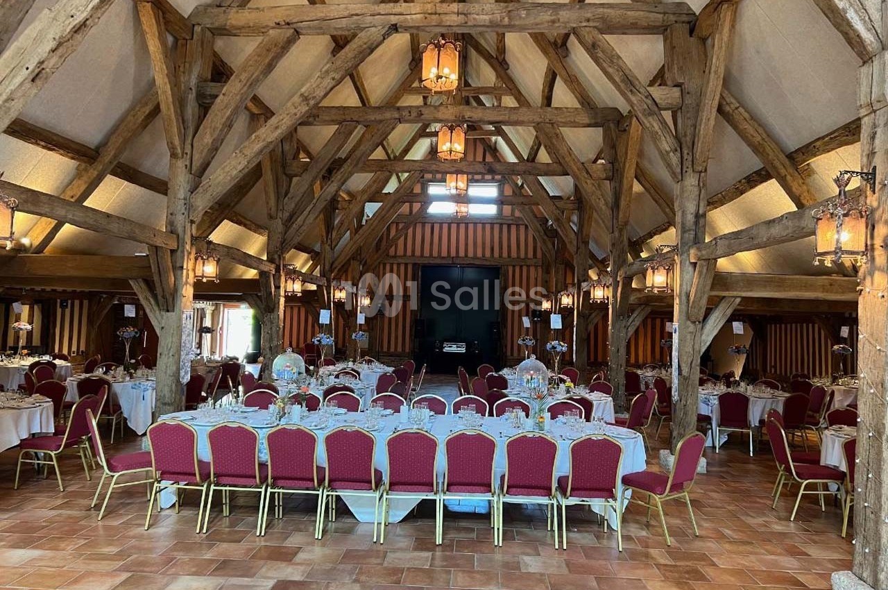 Salle de réception avec charpente en bois apparente, tables dressées et chaises rouges sur un sol en carrelage.