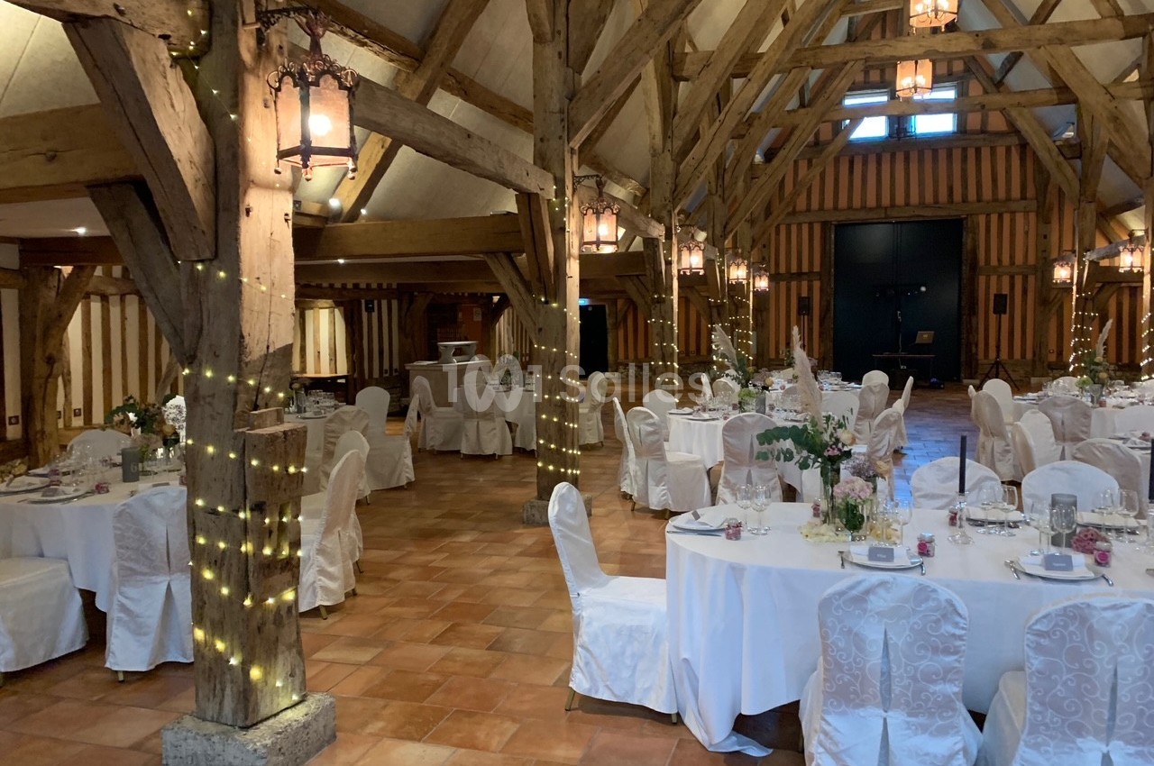 Salle de réception avec poutres apparentes, tables rondes dressées et chaises recouvertes de housses blanches.